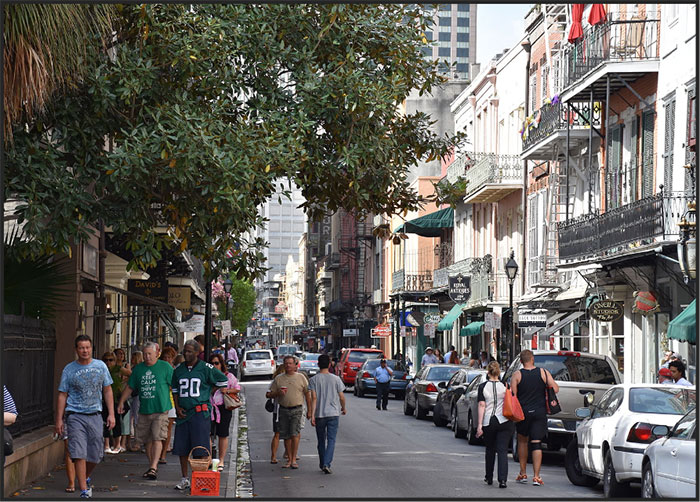 New Orleans street with pedastrians and cars shaded by trees and historic buildings with balconies.